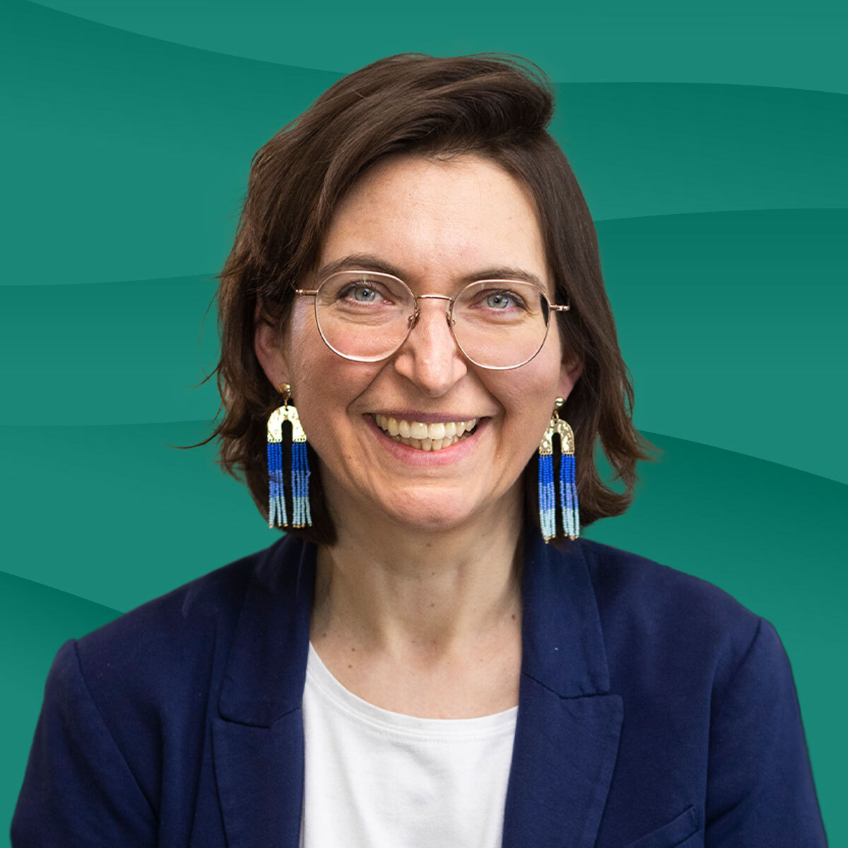 Porträt einer lächelnden Frau mit Brille und auffälligen Ohrringen, vor einem grünen Hintergrund.